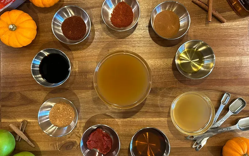 Ingredients for apple cider BBQ sauce in small bowls on a wooden board with cinnamon sticks and mini pumpkins