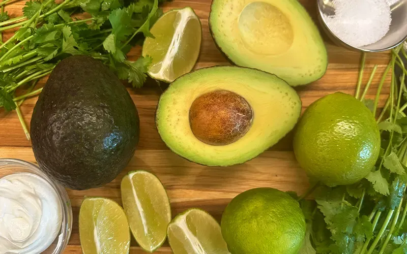 Halved avocados, limes, cilantro and sour cream on a wooden board