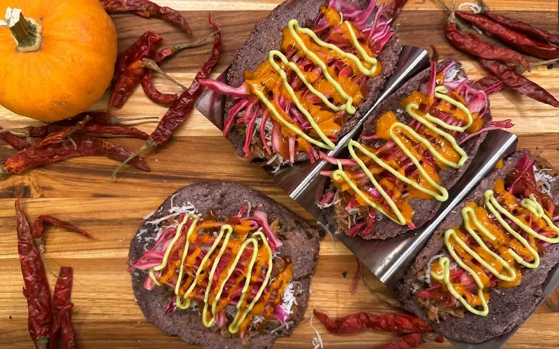 Pulled pork tacos on blue corn tortillas topped with purple cabbage and drizzled sauces, on a board with dried chiles and small pumpkins