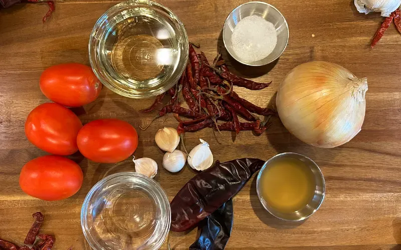 Tomatoes, onion, garlic, dried chiles and small bowls of liquid on a wooden board
